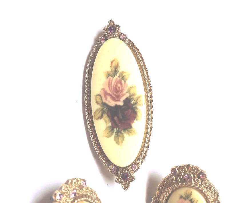 May include: An oval brooch and two earrings with a floral design. The brooch features a cream-colored oval with a painting of pink and burgundy roses. The brooch and earrings have a gold-colored metal frame with pink accents.