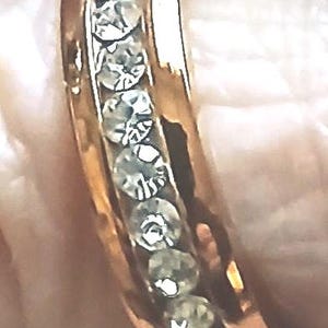 May include: A gold-colored ring with a row of clear, sparkling stones set along the top. The ring is shown close-up, highlighting the details of the stones and the band's polished finish. The ring is a piece of jewelry.