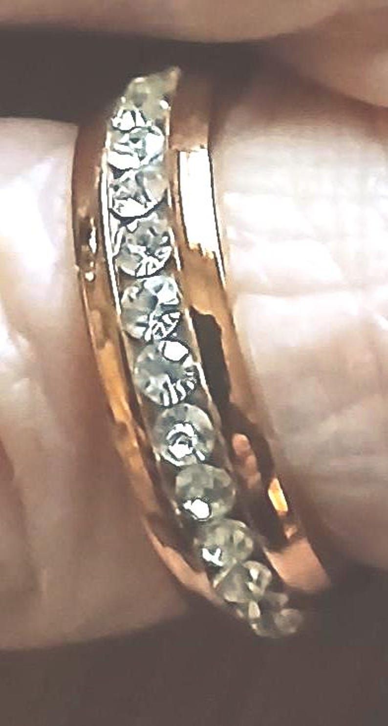 May include: A gold-colored ring with a row of clear, sparkling stones set along the top. The ring is shown close-up, highlighting the details of the stones and the band's polished finish. The ring is a piece of jewelry.