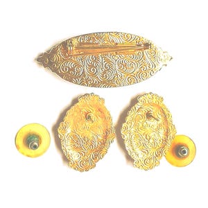 May include: A vintage jewelry set featuring a gold-toned brooch and matching earrings. The brooch is an elongated oval shape, and the earrings are oval-shaped. All pieces have an ornate, swirling pattern.