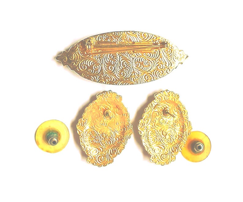 May include: A vintage jewelry set featuring a gold-toned brooch and matching earrings. The brooch is an elongated oval shape, and the earrings are oval-shaped. All pieces have an ornate, swirling pattern.