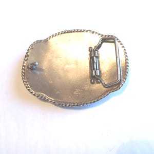 May include: Silver-toned oval belt buckle with a decorative rope-like border. The back features a hinged bar and a pin for securing the belt. The buckle is designed for use with a belt and is made of metal.