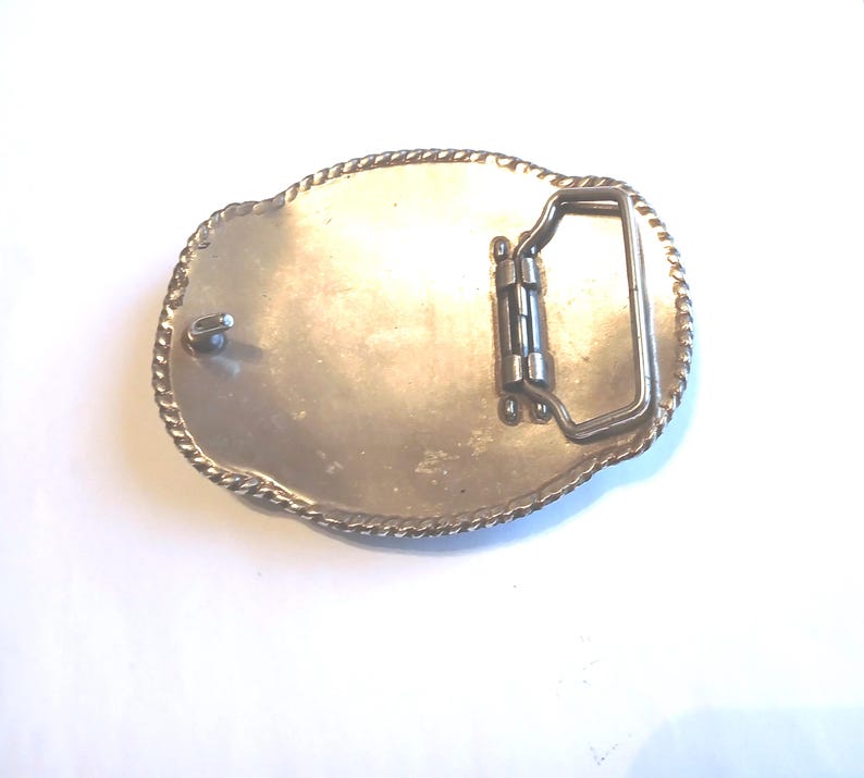 May include: Silver-toned oval belt buckle with a decorative rope-like border. The back features a hinged bar and a pin for securing the belt. The buckle is designed for use with a belt and is made of metal.