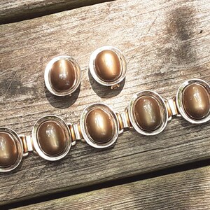 以下が含まれることがあります： リンクブレスレットとクリップ式イヤリングのジュエリーセット。各ピースには、光沢のあるシルバーカラーの金属フレームにセットされた、滑らかな楕円形のメタリックブロンズトーンのカボションがあしらわれています。ブレスレットは5つの楕円形のリンクで構成されています。