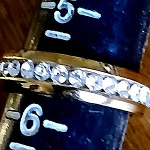 May include: A gold-colored ring with a row of clear, sparkling stones set along the top. The ring is positioned next to a ruler, with the number 6 visible. The ring's design is simple and elegant.