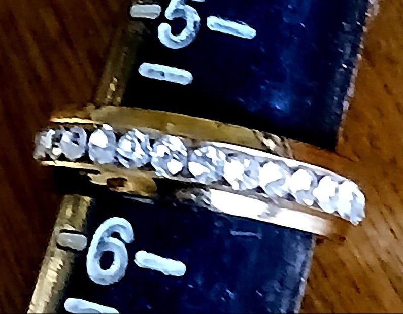 May include: A gold-colored ring with a row of clear, sparkling stones set along the top. The ring is positioned next to a ruler, with the number 6 visible. The ring's design is simple and elegant.