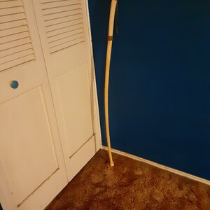 Op de afbeelding: Een lange, houten wandelstok met een bruine leren wikkeling aan de bovenkant. De stok leunt tegen een witte deur en een blauwe muur.