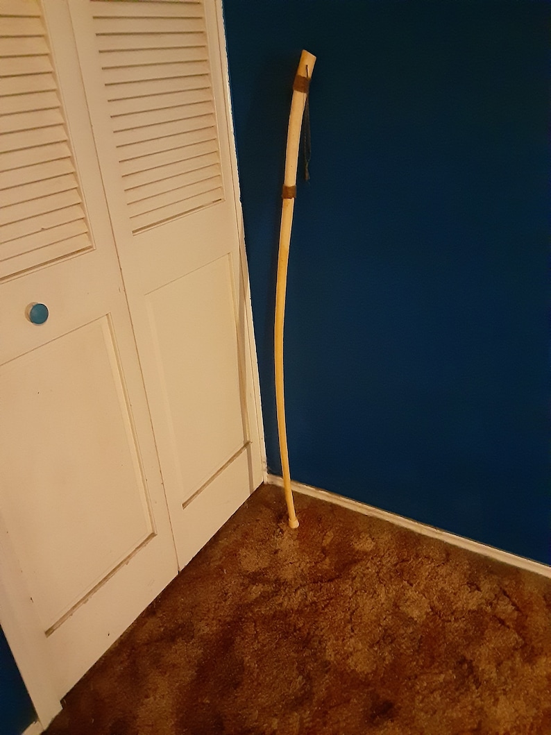 Op de afbeelding: Een lange, houten wandelstok met een bruine leren wikkeling aan de bovenkant. De stok leunt tegen een witte deur en een blauwe muur.