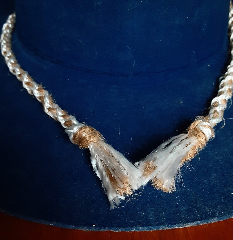 Puede incluir: Un collar de cuerda blanco y marr&oacute;n con borlas sobre un fondo azul.