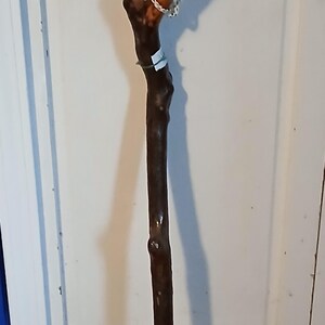 May include: A wooden walking stick with a brown, natural finish. The stick has a curved handle and a white tip. The stick is hanging on a white door with a blue and white rope.
