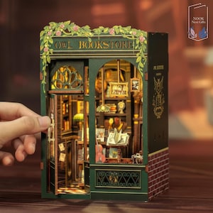 May include: Miniature green and brown Owl Bookstore model with the words "OWL BOOKSTORE" above the entrance. The model has an open door, interior lighting, and detailed bookshelves. The exterior features floral accents and a brick facade.