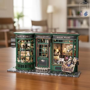 Puede incluir: Modelo en miniatura de una tienda de varitas mágicas con exterior verde y marrón. La tienda tiene múltiples ventanas que exhiben varios artículos. El letrero dice "MAGIC WAND" y "MAGICAL WAND SHOP". Un pequeño carro está al frente.