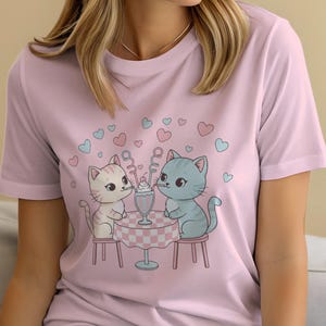 May include: Pink t-shirt featuring a cartoon illustration of two cats sharing a milkshake at a table. The cats are seated on stools, with hearts floating above them. The table has a pink and white checkered tablecloth.