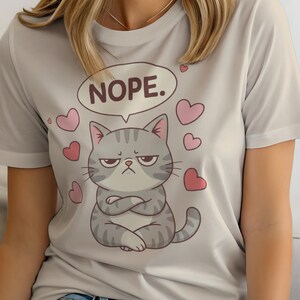 May include: A light grey t-shirt featuring a cartoon cat with a grumpy expression and crossed arms. The cat is gray with darker stripes and is surrounded by pink hearts. A speech bubble above the cat says "NOPE."