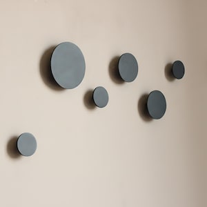 Puede incluir: Una colección de siete perchas de pared circulares de color gris oscuro de varios tamaños están montadas en una pared beige claro. Las perchas están dispuestas en un patrón asimétrico, ofreciendo una estética moderna y minimalista.