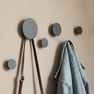 Può includere: Un set di sei ganci a parete grigi e circolari di varie dimensioni montati su una parete beige. Una giacca di jeans blu è appesa a uno dei ganci e una borsa bordeaux è appesa a un altro. I ganci offrono una soluzione di archiviazione moderna.