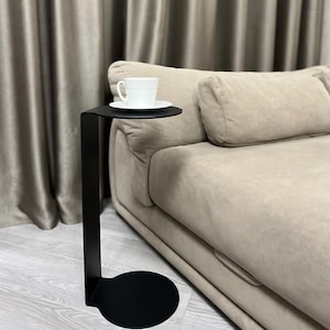 Puede incluir: Una mesa auxiliar de metal negro con base redonda y tapa circular. Una taza de té y un platillo blancos están sobre la mesa. La mesa está al lado de un sofá beige.