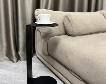 May include: A black metal side table with a round base and a circular top. A white teacup and saucer sit on the table's surface. The table is next to a beige sofa.