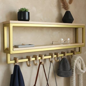 Può includere: Una moderna mensola da parete con struttura in metallo color oro e ripiani in legno naturale. La mensola contiene una piccola pianta in vaso, un vassoio e occhiali. Sotto, una fila di ganci tiene un cappotto, una borsa, un cappello e una sciarpa. Una soluzione di archiviazione elegante.