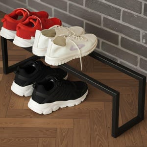 Puede incluir: Un zapatero de metal negro con tres pares de zapatillas deportivas. Las zapatillas son rojas, blancas y negras con suelas blancas. El zapatero está contra una pared de ladrillo gris y sobre un suelo de madera.