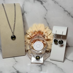 Könnte beinhalten: Ein Schmuckset mit Halskette, Armband und Ohrringen. Jedes Teil hat eine silberne Kette und einen runden, schwarz-gelben Anhänger. Der Schmuck wird auf weißen Ständern und einem transparenten Halter präsentiert.
