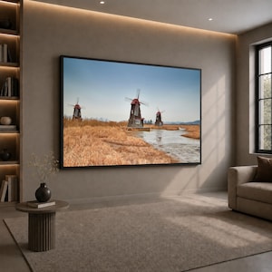 Puede incluir: Un gran cuadro rectangular de pared que muestra tres molinos de viento de madera oscura con detalles rojos, situados en un campo de hierba seca de color marrón dorado con un pequeño arroyo bajo un cielo azul claro, montado en una pared interior.