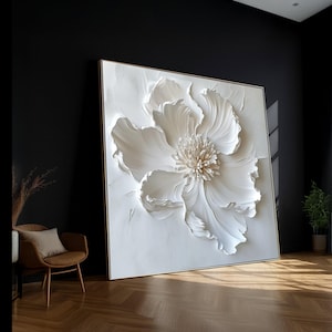 Può includere: Grande opera d'arte quadrata con un design floreale bianco strutturato. L'opera è incorniciata in una cornice dorata e appoggiata a una parete nera. I petali e il centro del fiore presentano dettagli intricati.