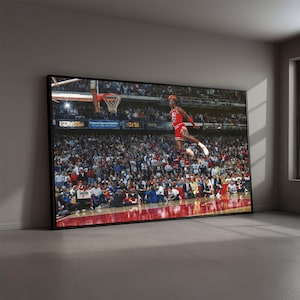 Könnte beinhalten: Gerahmter Druck eines Basketballspielers beim Dunk, in einem roten Trikot und Shorts. Der Hintergrund zeigt ein überfülltes Stadion mit Zuschauern. Der Druck wird in einem Raum mit grauer Wand und Fenster ausgestellt.