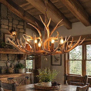 May include: A rustic antler chandelier with multiple candle-style lights. The chandelier is made of natural-looking antlers and has a warm, inviting glow. The fixture hangs from a chain and is set against a backdrop of wooden beams and a stone wall.