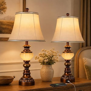 May include: Two matching bronze-colored table lamps with cream-colored shades. Each lamp has a decorative base with a ribbed glass section and a fabric shade trimmed with black. The lamps are placed on a wooden surface.