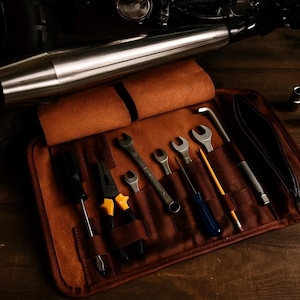 Puede incluir: Un rollo de herramientas de cuero marr&oacute;n con varias herramientas manuales, incluyendo llaves, destornilladores y alicates. Un tubo de escape de motocicleta es visible en el fondo. Las herramientas est&aacute;n dispuestas sobre una superficie de madera.