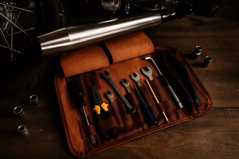 Puede incluir: Un rollo de herramientas de cuero marr&oacute;n con varias herramientas manuales, incluyendo llaves, destornilladores y alicates. Un tubo de escape de motocicleta es visible en el fondo. Las herramientas est&aacute;n dispuestas sobre una superficie de madera.