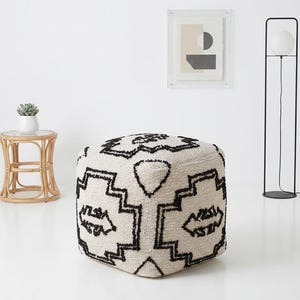 May include: A square, off-white ottoman with a geometric black pattern. The design features angular shapes and lines. A small, round side table with a glass top and a succulent plant in a white pot are in the background.