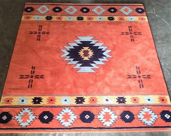 Puede incluir: Una alfombra rectangular con un fondo naranja vibrante y un motivo central de diamante geométrico en azul oscuro, azul claro y amarillo. La alfombra incluye patrones de borde horizontales con formas repetitivas y cuatro símbolos en cruz.