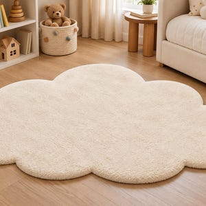 May include: A cream-colored, plush area rug shaped like a cloud with rounded, scalloped edges. The rug features a soft, textured surface and is laid flat on a wooden floor, showcasing a simple, organic silhouette without any additional patterns.
