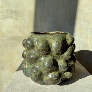 Può includere: Un vaso in ceramica fatto a mano con una superficie strutturata unica. Il vaso presenta un design organico e a bolle con una finitura lucida e macchiata nei toni del verde, del marrone e del rosso. L'interno è scuro, in contrasto con l'esterno.