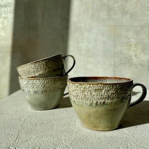 Pode incluir: Três canecas de cerâmica empilhadas com uma superfície texturizada e salpicada. As canecas têm um gradiente de cores, passando do verde claro na base para o castanho na borda. Cada caneca tem uma alça arredondada.