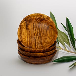 Peut inclure: Une pile de petits bols ronds en bois, de couleur brun chaud et présentant des motifs de grain de bois uniques. Les bols sont en bois d'olivier et sont présentés sur une surface blanche, avec des feuilles d'olivier vertes en arrière-plan.