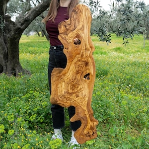 May include: A large, irregularly shaped wooden slab with a natural edge. The slab is a warm, light brown colour with darker brown grain patterns and several knot holes. The wood appears to be olive wood. The slab is held upright in a grassy field.