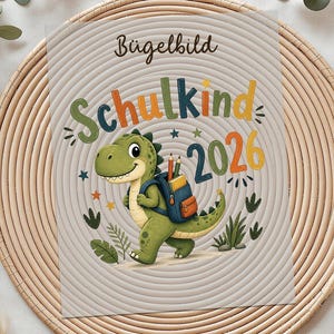 Könnte beinhalten: Ein Bügelbild mit einem freundlichen grünen Dinosaurier mit Rucksack, dem Text "Schulkind 2026" und dekorativen Elementen. Das Bügelbild liegt auf einem geflochtenen Untersetzer mit Dekoration.