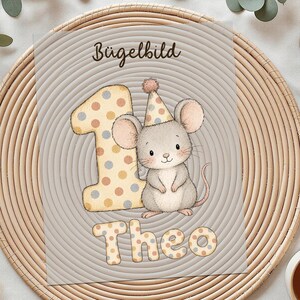 Könnte beinhalten: Ein rechteckiger Bügelbild-Transferbogen mit einer grauen Maus mit Partyhut, einer großen gepunkteten Eins und dem Namen Theo in passender gepunkteter Schrift. Der Text Bügelbild ist oben auf dem Design aufgedruckt.