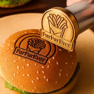 Può includere: Un panino con semi di sesamo con un logo stampato "FurFurEver" e un timbro dorato con lo stesso logo. Il timbro viene premuto sul panino. Il logo presenta un'immagine stilizzata di patatine fritte.