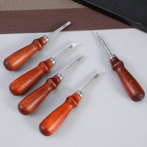 Pode incluir: Cinco ferramentas de artesanato em couro com cabos de madeira e lâminas de metal prateado. As ferramentas estão dispostas sobre uma superfície branca, mostrando sua madeira polida e construção metálica. Os cabos são de uma rica cor castanho-avermelhada.