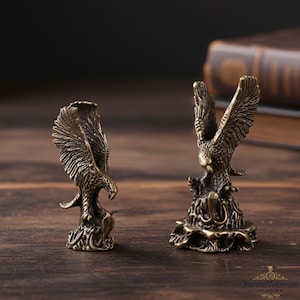 Puede incluir: Dos figuras de águila de bronce con las alas extendidas. Las esculturas detalladas presentan intrincados diseños de plumas y están montadas sobre bases decorativas. Las figuras se exhiben sobre una superficie de madera, mostrando la artesanía.