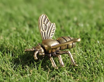 Peut inclure: Une figurine de bee en laiton détaillée, avec des détails complexes sur les ailes et le corps. L'abeille est posée sur de l'herbe verte, mettant en valeur ses pattes, ses antennes et son dard. L'objet est décoratif.