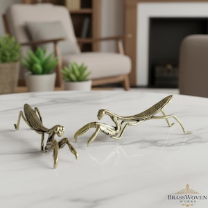 Puede incluir: Dos esculturas de mantis religiosas de latón sobre una mesa de mármol blanco. Las esculturas presentan diseños detallados de alas y patas. Las mantis están en diferentes poses, destacando sus formas únicas. Esculturas decorativas y artísticas.