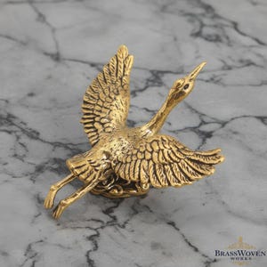 May include: A detailed, gold-coloured brass sculpture of a bird in flight. The bird has outstretched wings and detailed feathering. The sculpture is set against a marble-effect background, with the Brass Woven Works logo in the bottom right corner.