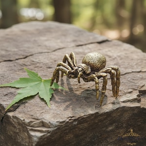 Può includere: Una scultura dettagliata di ragno in ottone con otto zampe e un corpo strutturato, appoggiata su una superficie di pietra. Una foglia verde è accanto al ragno. La scultura è un oggetto decorativo.