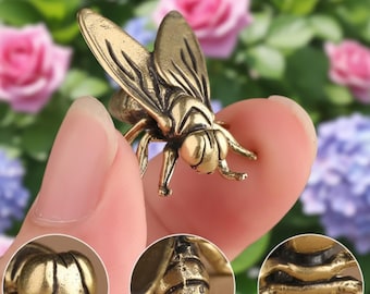 May include: A detailed brass fly figurine with intricate wing and body details. The insect sculpture is a golden color and appears to be a decorative item. The background features blurred pink and purple flowers.