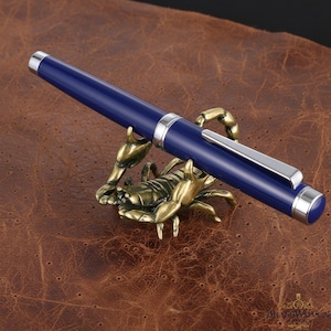 May include: A navy blue pen with silver accents rests on a bronze scorpion-shaped pen holder. The pen holder is detailed with intricate textures and sits on a brown leather surface.